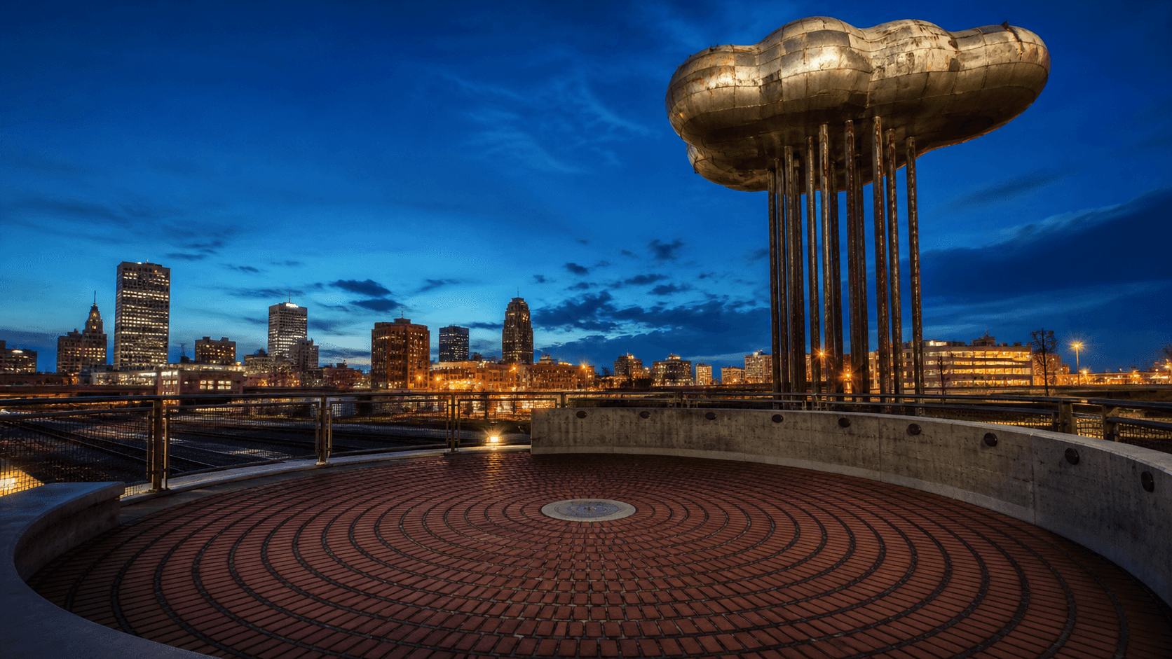 Editorial illustration of the brick acoustic circle and steel public art above downtown Tulsa railroad tracks at blue hour.